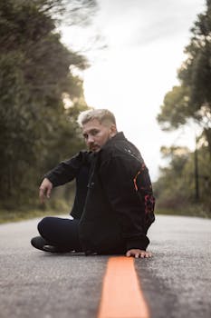 Trendy young man with blonde hair sitting on a road in Breves, Brazil, in casual fashion.
